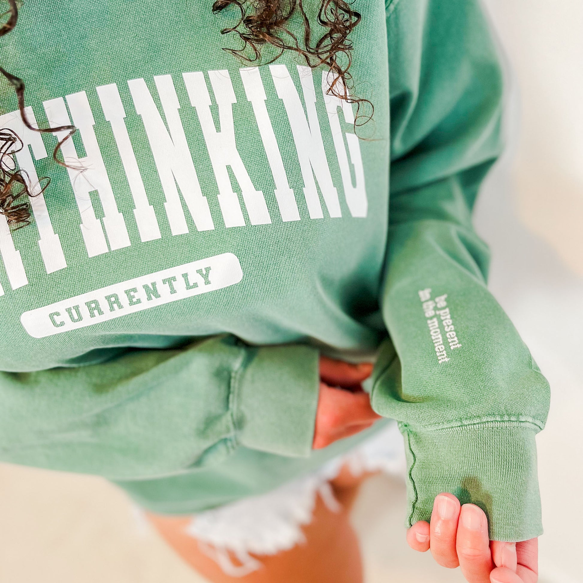 The model is wearing a light green vintage-washed crewneck sweatshirt with the words "CURRENTLY OVERTHINKING" printed in capitalized athletic-style lettering. This design is placed in the centre of the sweater, 4" below the collar. On the bottom left sleeve are the embroidered words: be present in the moment. 